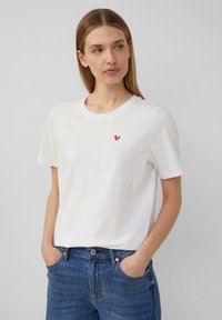 T-shirt en coton blanc avec un petit design de cœur rouge sur la poitrine. Manches courtes et style col rond classique. Porté avec un jean en denim bleu.