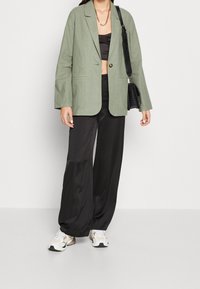 Světle zelený lněný blejzr přes černý crop top, v kombinaci s černými širokými kalhotami a bílými teniskami s béžovými a zelenými akcenty. Černá taška.