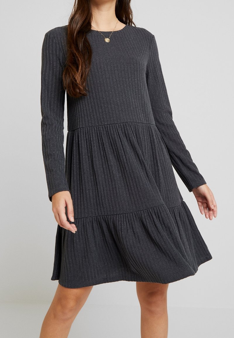 Femme portant une robe tricotée côtelée gris foncé à manches longues et une jupe à volants, debout devant un fond clair uni.