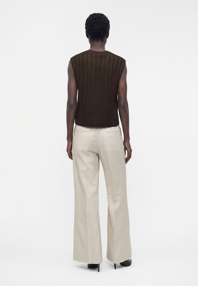 Personne aux cheveux courts et bouclés portant un haut en maille marron foncé sans manches et un pantalon beige à jambes larges, debout de dos sur un fond blanc.