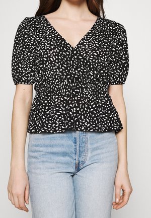 Kvinde iført sort-hvid bluse med abstrakt mønster, V-udskæring, pufærmer og lyseblå højtsiddende jeans.