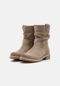 Bottes chelsea en daim beige avec un design moelleux, semelle en caoutchouc et couture ton sur ton. Présentent une sangle sur le côté et un bout arrondi.