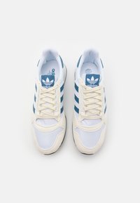 Tenisky Adidas s bílým síťovaným svrškem, modrými třemi pruhy a krémovými akcenty. Mají texturovanou podešev a zavazování na tkaničky.