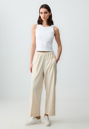 Top bianco senza maniche abbinato a pantaloni larghi beige chiaro con cintura elastica e tasche laterali. Le scarpe sono sneaker bianche.