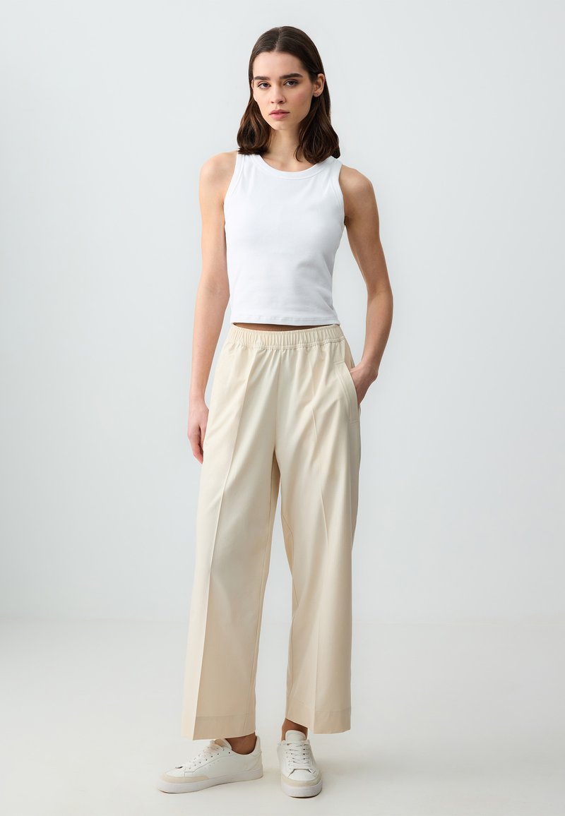 Top bianco senza maniche abbinato a pantaloni larghi beige chiaro con cintura elastica e tasche laterali. Le scarpe sono sneaker bianche.