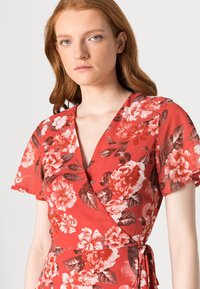 Vestido envolvente floral em coral com mangas curtas. Possui um decote em V profundo e uma cintura amarrada. O design inclui grandes flores brancas e vermelhas e folhas verdes.