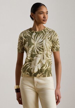 Vrouw draagt een kort groen-beige top met tropisch bladdessin, gebroken-witte broek en twee armbandjes aan haar linkerpols.