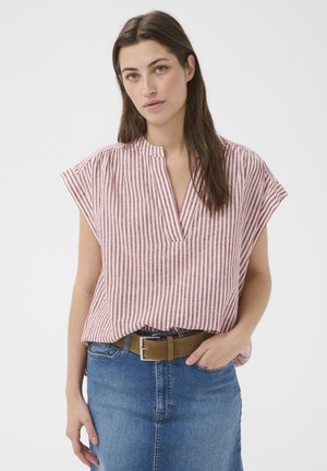 Femme aux longs cheveux bruns portant un chemisier sans manches rouge et blanc rayé, rentré dans un jean bleu avec une ceinture marron.