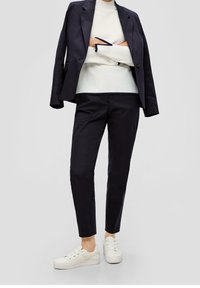 Person som bär en marinblå kavaj över axlarna, marinblå skräddarsydda byxor, vit tröja och vita sneakers, står med armarna i kors.