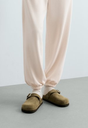 Pieds portant un pantalon de survêtement beige clair, des chaussettes blanches et des sabots en daim vert olive avec une boucle latérale, debout sur un sol gris.