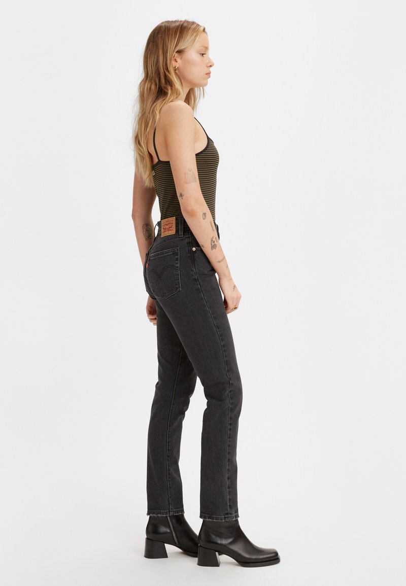 Schwarze Denim-Jeans mit schlanker Passform, rohem Saum und klassischen Gesäßtaschen. Kombiniert mit einem gestreiften Tanktop und schwarzen Knöchelstiefeln.