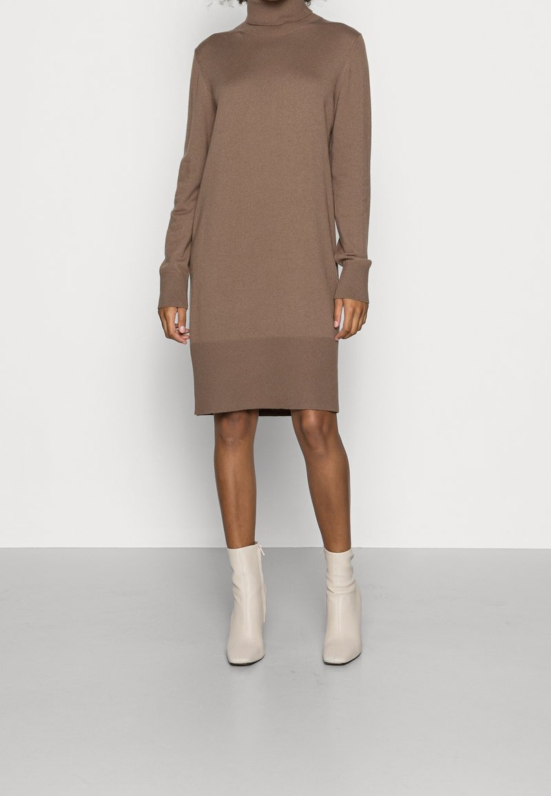 Femme portant une robe pull marron à col roulé et longueur genoux, avec des bottines écrues, posant devant un fond gris clair uni.
