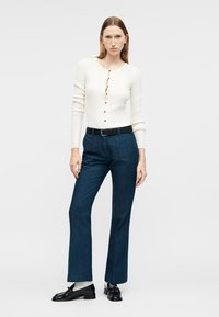 Γυναίκα που φοράει εφαρμοστό λευκό πλεκτό με κουμπιά, τζιν ψηλόμεσο σκούρο μπλε, μαύρη ζώνη, λευκές κάλτσες και μαύρα μοκασίνια, στέκεται μπροστά σε απλό φόντο.