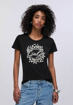 Donna con capelli ricci indossa una maglietta nera con logo floreale e jeans azzurri strutturati, posando davanti a uno sfondo grigio chiaro.