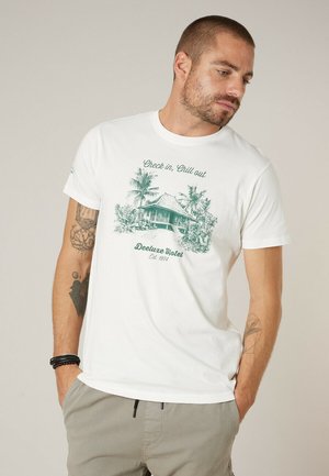 Homme aux cheveux courts et barbe portant un t-shirt blanc avec un motif de hutte tropicale verte et le texte "Check in, Chill out Deeluxe Hotel Est. 1974."