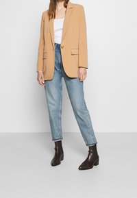 Beige blazer met gestructureerde schouders, twee voorzakken, sluiting met één knop; gecombineerd met lichtblauwe tapering jeans en donkerbruine enkellaarsjes.