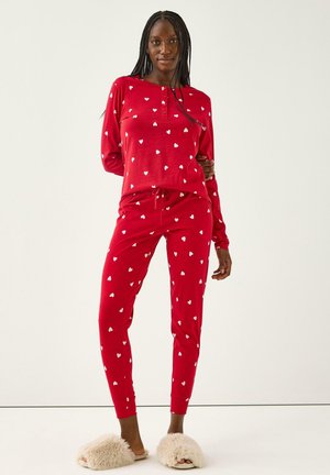 Set di pigiama rosso con stampe a cuori bianchi, composto da una maglietta a maniche lunghe e pantaloni affusolati con vita elastica e cordoncino. Pantofole in morbido materiale.