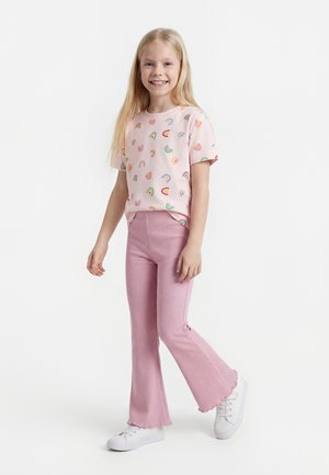 Ragazza giovane che sorride, in piedi con un piede avanzato, indossa una maglietta rosa con motivo arcobaleno, pantaloni rosa a zampa e scarpe da ginnastica bianche.