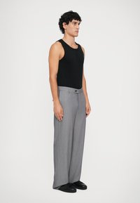 Man met krullend haar staat zijwaarts gekeerd in een zwart mouwloos shirt, grijze wijde broek en zwarte schoenen tegen een effen achtergrond.