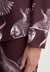 Bordeaux pyjama set met witte uil- en vogelpatronen. Zachte stof met piping op de manchetten. Close-up van de mouw en handdetails.