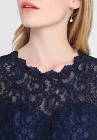 Gros plan d'une femme portant un haut en dentelle bleu marine avec des bords festonnés et de longues boucles d'oreilles pendantes ornées de pierres rondes transparentes.