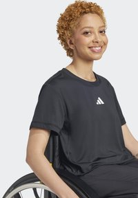 Juodas sportinis marškinėliai su trumpomis rankovėmis, su baltu Adidas logotipu ant krūtinės. Lygus paviršius, nešiojamas asmens vežimėlyje.