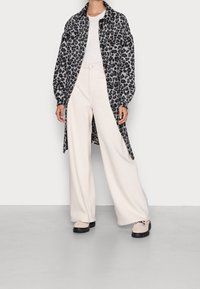 Veste-chemise longue à imprimé léopard noir et gris, haut blanc cintré, pantalon large crème et chaussures beiges avec des accents noirs.