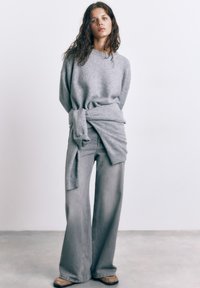 Kvinde med krøllet hår iført grå, oversized sweater og viddebukser, stående mod en ensfarvet hvid væg og betongulv.