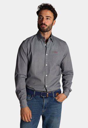 Donkere grijze overhemd met een klein wit stippenpatroon, voorzien van een puntsluiting en contrasterende knopen. Gecombineerd met jeans en een blauwe riem.