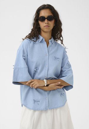 Femme aux longs cheveux bouclés portant des lunettes de soleil noires et une chemise bleu clair à boutons avec de petits détails en forme de nœud, debout les bras croisés.