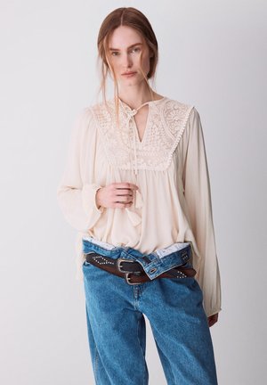 Mujer que lleva una blusa de encaje color crema y vaqueros azules con dos cinturones marrones atados alrededor de la cintura, de pie frente a un fondo liso.