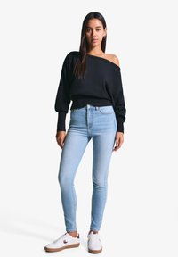 Crna majica bez rukava s rebrastim rubom, uparena sa svijetloplavim uskim trapericama i bijelim tenisicama s smeškastim akcentima.