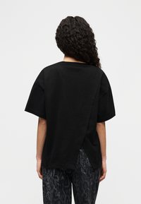 Μαύρο υπερμεγέθες μπλουζάκι με κοντά μανίκια, με λεπτομέρεια στη πλάτη και κατασκευασμένο από απαλό βαμβακερό ύφασμα. Φορεμένο με σκούρα τζιν με σχέδια.