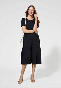 Czarna, casualowa sukienka z krótkimi rękawami, z falbanami, okrągłym dekoltem, miękką fakturą, w połączeniu z beżowymi sandałami na słupku i lekką torebką.