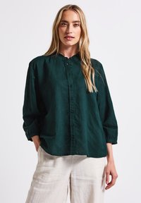 Dark green collared shirt made of lightweight fabric, featuring a button placket, rolled sleeves, and a relaxed fit. Paired with light linen trousers.