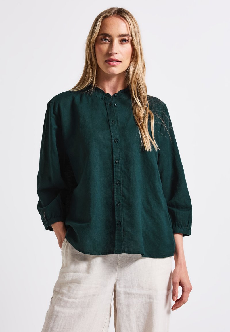 Dark green collared shirt made of lightweight fabric, featuring a button placket, rolled sleeves, and a relaxed fit. Paired with light linen trousers.