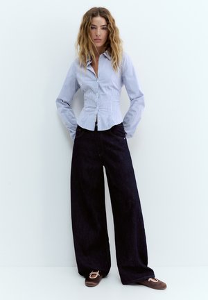 Hölgy hullámos hajjal, testhezálló, kék csíkos inggel, széles szárú fekete nadrággal és barna balettcipővel, egy fehér fal előtt állva.