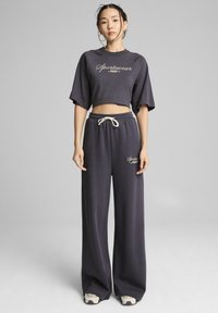 Completo coordinato grigio scuro composto da una maglietta corta a maniche corte e pantaloni a gamba larga con cintura elastica e un cordino. Dettaglio del logo presente.