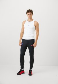 Hvid tanktop med rund halsudskæring, sammen med sorte ribbede joggers med blå og røde detaljer. Sorte træningssko fuldender looket.