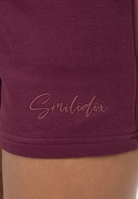 Maronin väriset puuvillashortsit, joissa on sileä pinta, ommeltu helma ja brändinimi "Smilodox" nostettuna vaaleanruskeana kirjaimena.