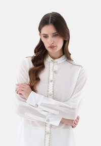 White button-up blouse with sheer long sleeves, gold stitching along the placket, and decorative buttons on the cuffs and front.