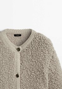 Cardigan beige texturé avec une surface douce et duveteuse. Présente un col rond, une fermeture à boutons, et des finitions côtelées au niveau du col.