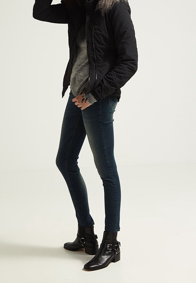 Personne portant un jean foncé ajusté, des bottines noires à boucles, un pull gris et une veste noire avec une capuche bordée de fourrure.