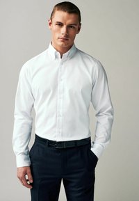 Camisa blanca de botones con cuello extendido, mangas largas y una textura de tela suave, combinada con pantalones oscuros ajustados y un cinturón negro.