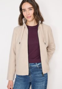 Donna che indossa una giacca in pile beige con zip sopra una maglietta viola e jeans blu, in piedi contro uno sfondo bianco semplice.