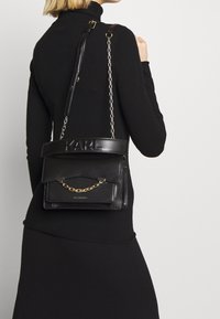 Donna con abito a collo alto nero che porta una borsa a tracolla in pelle nera di Karl Lagerfeld con catena e tracolla con logo in rilievo.