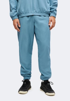 Mann trägt hellblaue Sweatpants mit elastischem Bund, passende Zip-up-Jacke, weiße Socken und schwarze Sportschuhe, steht mit entspannten Händen.