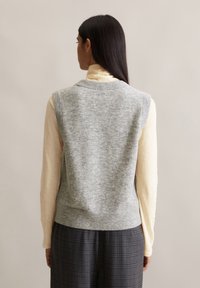 Un pull gris sans manches à tricot texturé, porté sur un haut à manches longues de couleur jaune clair. Le modèle est tourné le dos, avec une jupe à carreaux sombre visible.