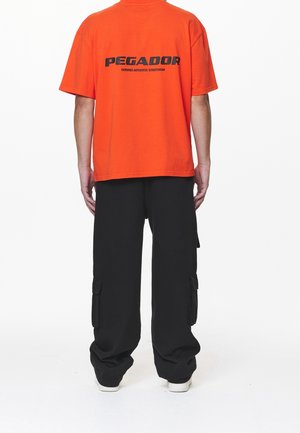 Orange kortærmet t-shirt med sort tekst på ryggen, parret med sorte cargo-bukser med sidelommer og en løs pasform.