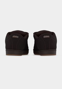 Ζευγάρι μαύρα παπούτσια Etnies, απεικονισμένα από πίσω, με επενδεδυμένους γιακάδες και καφέ εξωτερικές σόλες σε απλό λευκό φόντο.